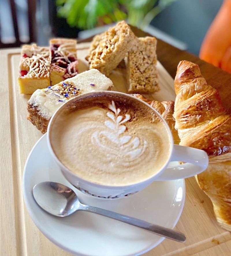 Cappuccino with latte art served alongside a croissant, flapjack, and assorted cake slices