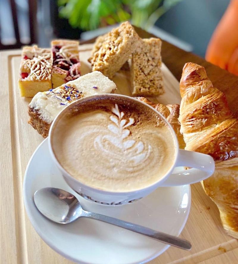 Cappuccino with latte art served alongside a croissant, flapjack, and assorted cake slices