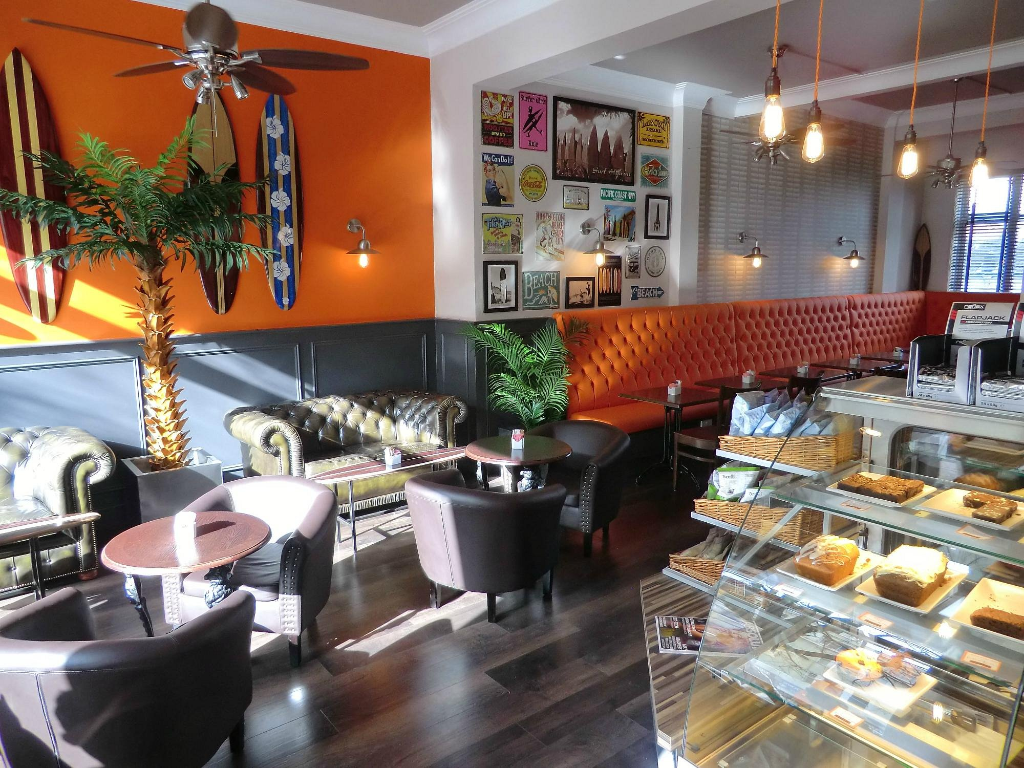 Modern café interior with leather armchairs and sofas, tropical plants, and orange feature wall decorated with surfboards. A long orange button-tufted bench lines the far wall under framed posters. A glass counter displays cakes in the foreground.